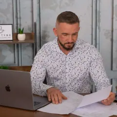 Mężczyzna w koszuli w kwiaty przegląda dokumenty przy biurku z laptopem i kawą.
