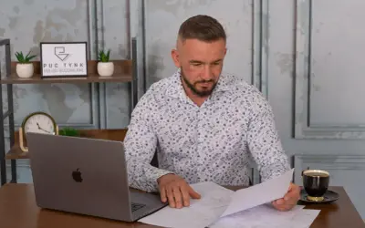 Mężczyzna w koszuli w kwiaty przegląda dokumenty przy biurku z laptopem i kawą.