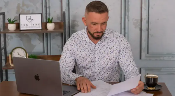 Mężczyzna w koszuli w kwiaty przegląda dokumenty przy biurku z laptopem i kawą.